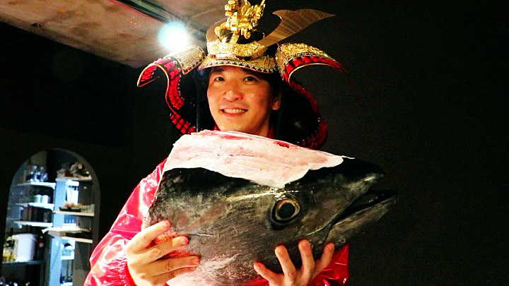 The host in a samurai helmet holding the tuna head